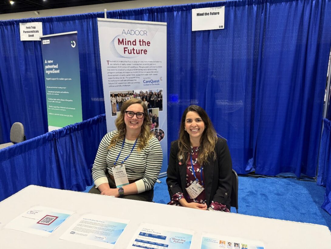 Two women smiling at a conference booth with a banner reading 