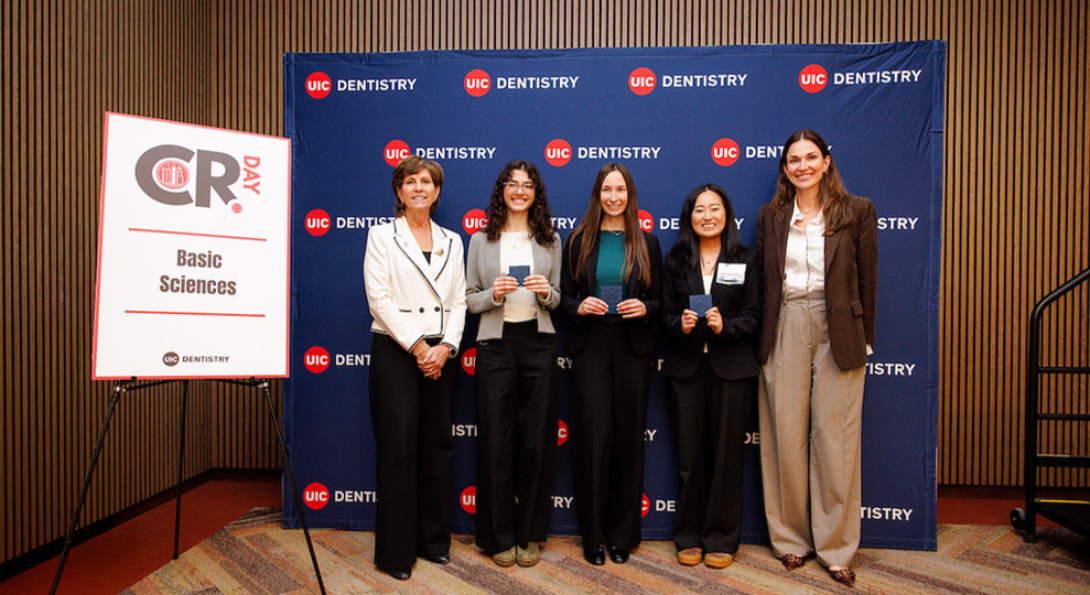 Reis-Havlat's lab team with Dr. Russon and Dean Rowan smiling in front of a UIC Dentistry backdrop. Two hold certificates, wearing professional attire, conveying a sense of achievement and pride.