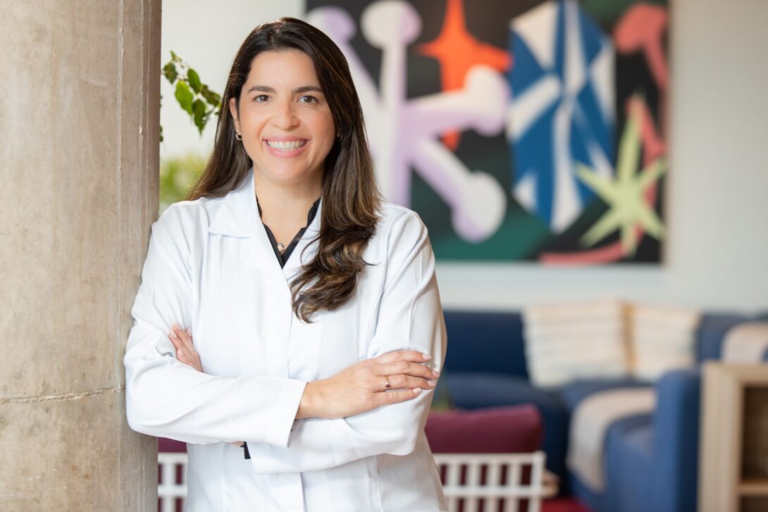 Dr. Mariana Reis – Havlat in a white lab coat smiles confidently with arms crossed, standing in a bright room with abstract art on the wall and blue furniture.
