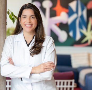Dr. Mariana Reis – Havlat in a white lab coat smiles confidently with arms crossed, standing in a bright room with abstract art on the wall and blue furniture. 