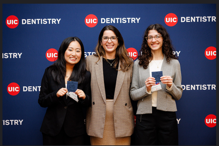 Reis-Havlat and her lab team smiling in front of a UIC Dentistry backdrop. Two hold certificates, wearing professional attire, conveying a sense of achievement and pride.